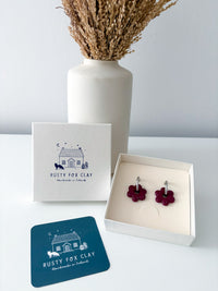 Rusty Fox Clay handmade earrings in a deep burgundy flower shape, displayed in an open cream-colored gift box next to the brand's white packaging box and a teal brand card, with a white ceramic vase of dried grass in the background.