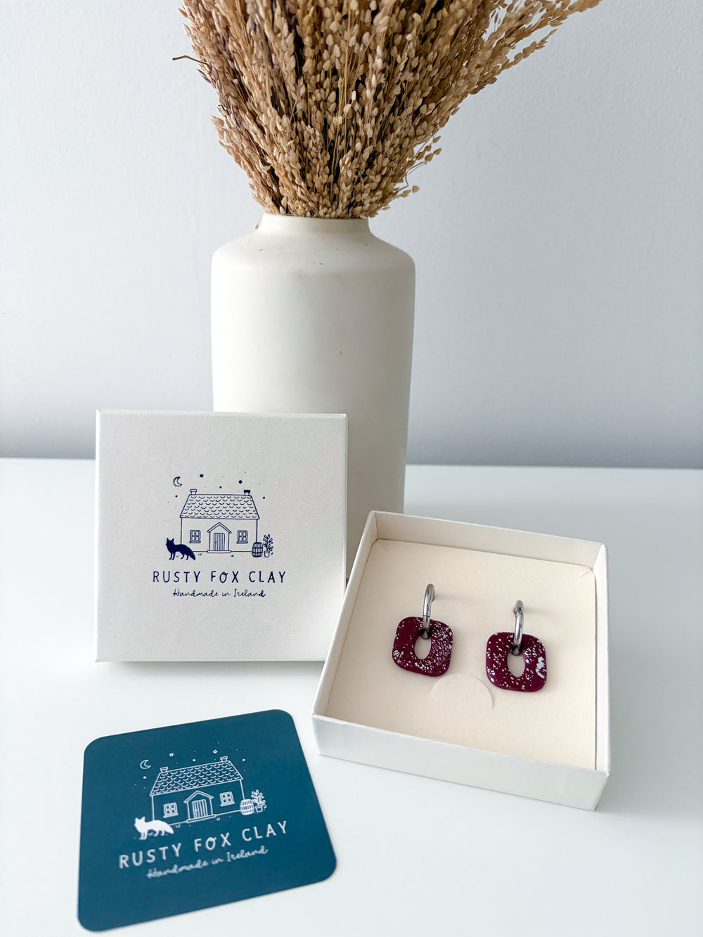 Rusty Fox Clay handmade earrings in a white gift box, displayed with the brand's logo box, a blue card, and a white vase with dried grass on a white surface.