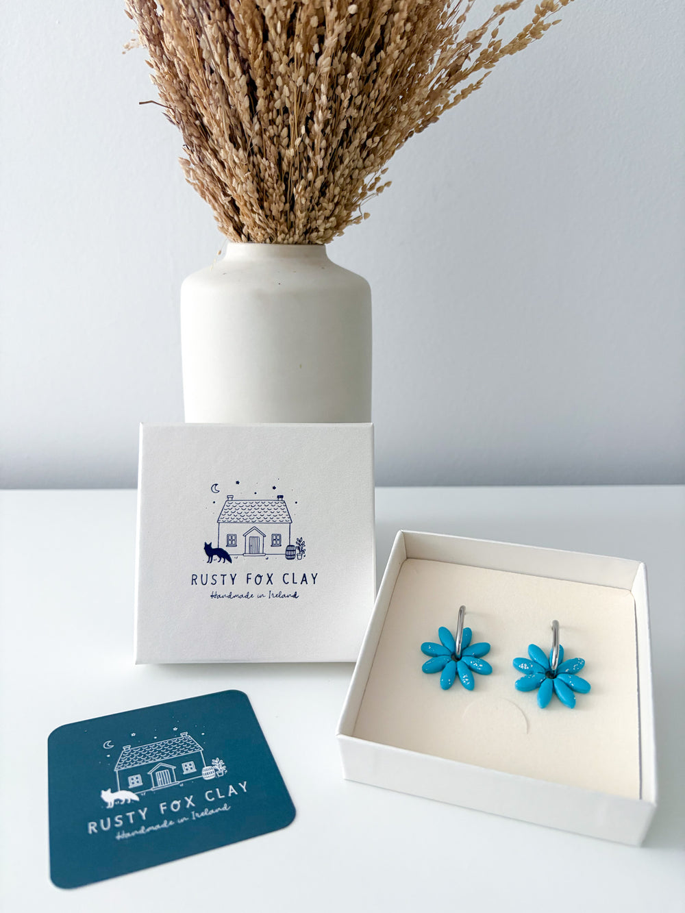 Blue flower-shaped clay earrings displayed in an open white gift box next to the Rusty Fox Clay branded packaging and a teal card, with a white vase of dried grass in the background.