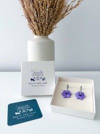 A pair of silver and purple-toned flower-shaped earrings displayed in an open white gift box, alongside the brand card, a blue coaster, and a white vase with dried grass on a light gray background.