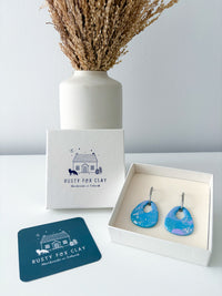 Blue and purple drop earrings displayed in an open white gift box next to the brand's white packaging box and a dark blue coasters, with a white vase of dried grass in the background.