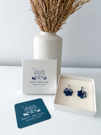 A pair of blue flower-shaped earrings displayed in an open white gift box, with the brand card 'Rusty Fox Clay' standing behind it and a matching teal card in front, set on a white surface with a white vase of dried grass against a light gray wall.