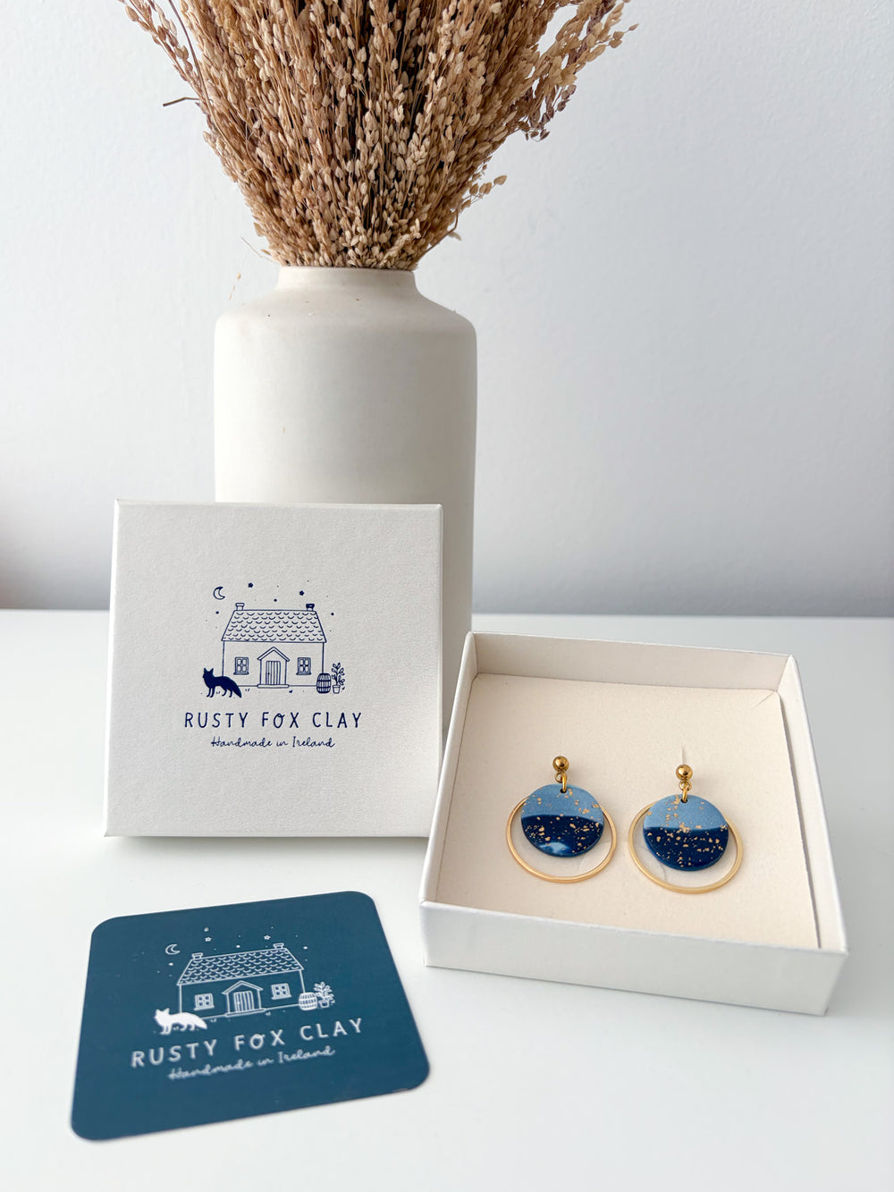 A pair of circle dangle earrings with blue and aqua starry sky designs and gold-tone borders, displayed in an open cream-colored gift box next to the brand's white packaging box and a dark blue branded card on a white surface.