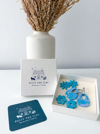 Blue and purple set of polymer clay earrings  displayed in a white gift box next to a branded Rusty Fox Clay box and card, with dried grass in a white vase in the background.