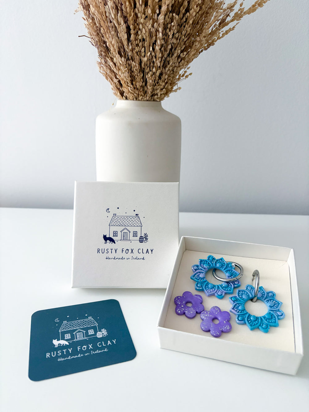 Three handmade clay charms in blue, purple, and teal are displayed in an open white gift box next to a branded white box and a blue coaster. A white ceramic vase with dried grass sits in the background against a light gray wall.
