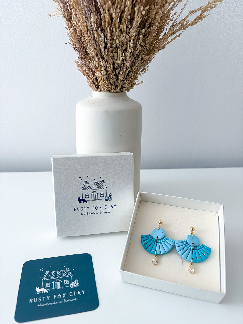 Turquoise fan-shaped clay earrings with gold hooks and small pearl drops, displayed in an open white gift box next to the Rusty Fox Clay branded box and a teal branded card, with a white vase of dried grass in the background.