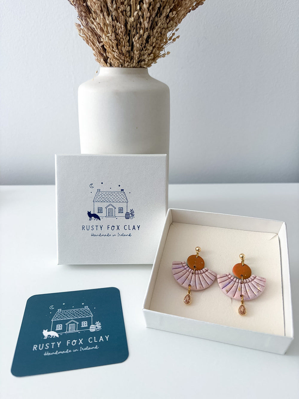A pair of Rusty Fox Clay handmade earrings displayed in an open white gift box next to the branded packaging and a blue card, with a white vase of dried grass in the background.