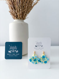 A pair of Rusty Fox Clay handmade earrings displayed on a white card, with a dark blue brand card and a white vase of dried grass in the background.