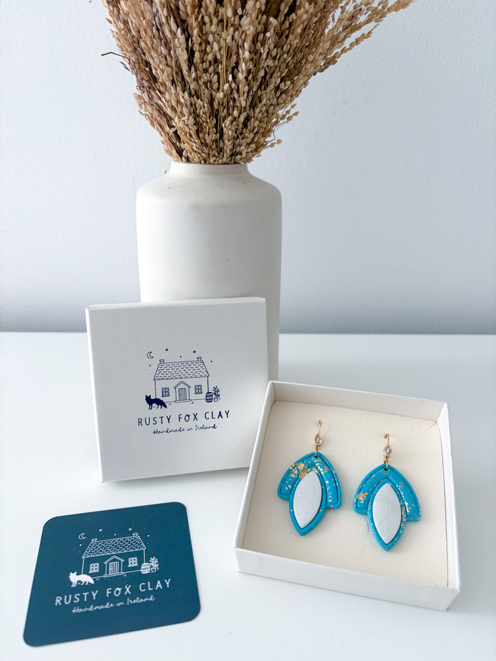 Turquoise and white crescent-shaped earrings displayed in an open white gift box next to the Rusty Fox Clay brand box and a branded card, with a white vase of dried grass in the background.