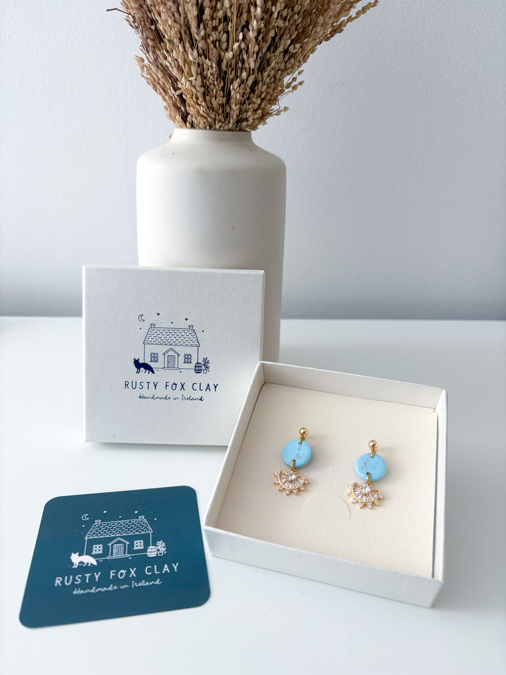A pair of Rusty Fox Clay handmade earrings displayed in an open white gift box, with the branded outer box and a dark teal card placed beside it on a white surface. A white ceramic vase with dried grass stands in the background against a light gray wall.