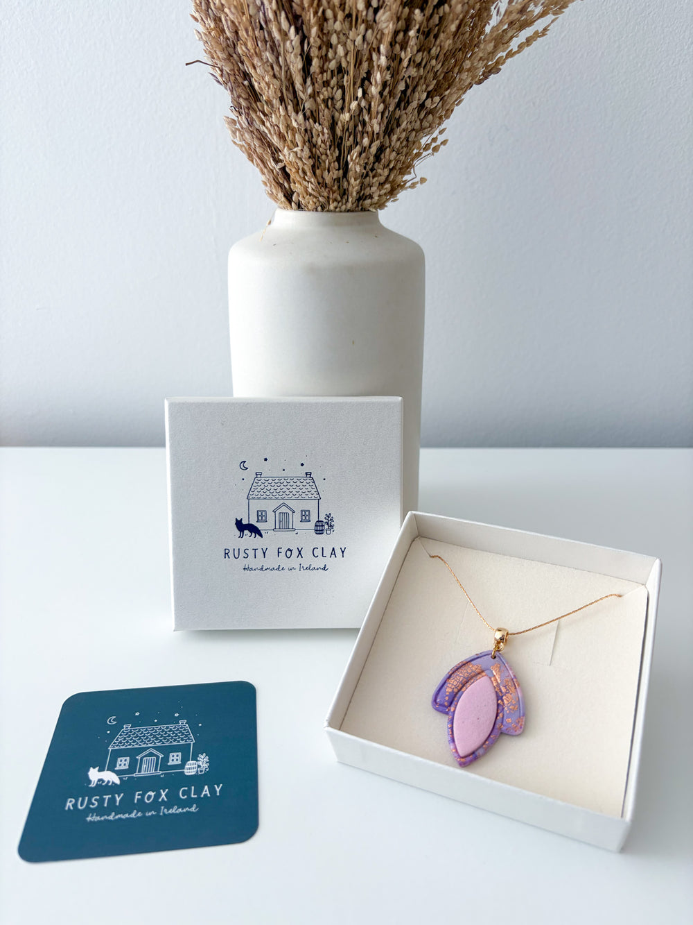 A handmade pendant necklace by Rusty Fox Clay, featuring a purple and pink textured clay heart on a gold chain, displayed in an open white gift box next to its branded packaging and a teal card.
