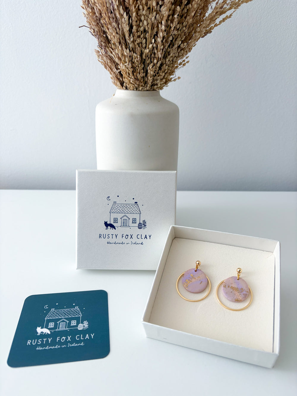 Rusty Fox Clay handmade earrings with round lavender and gold marbled pendants in a white box, shown with the brand's gift box and a blue card on a white surface.