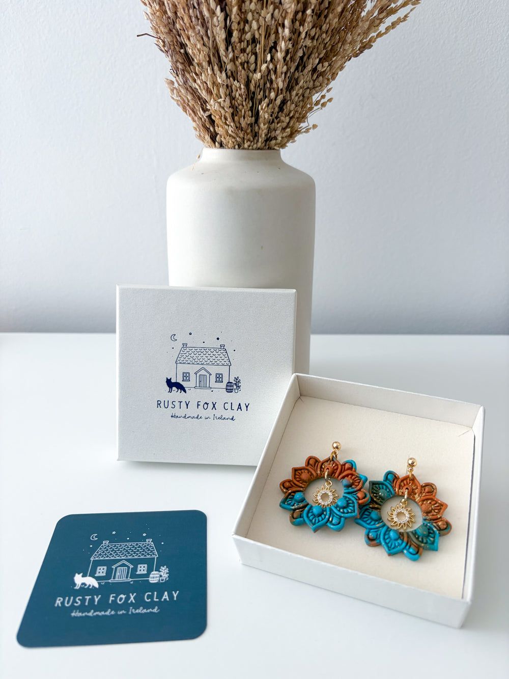 A pair of handmade clay earrings by Rusty Fox Clay, displayed in an open white gift box next to its branded packaging and a blue card, with a white vase of dried grass in the background.
