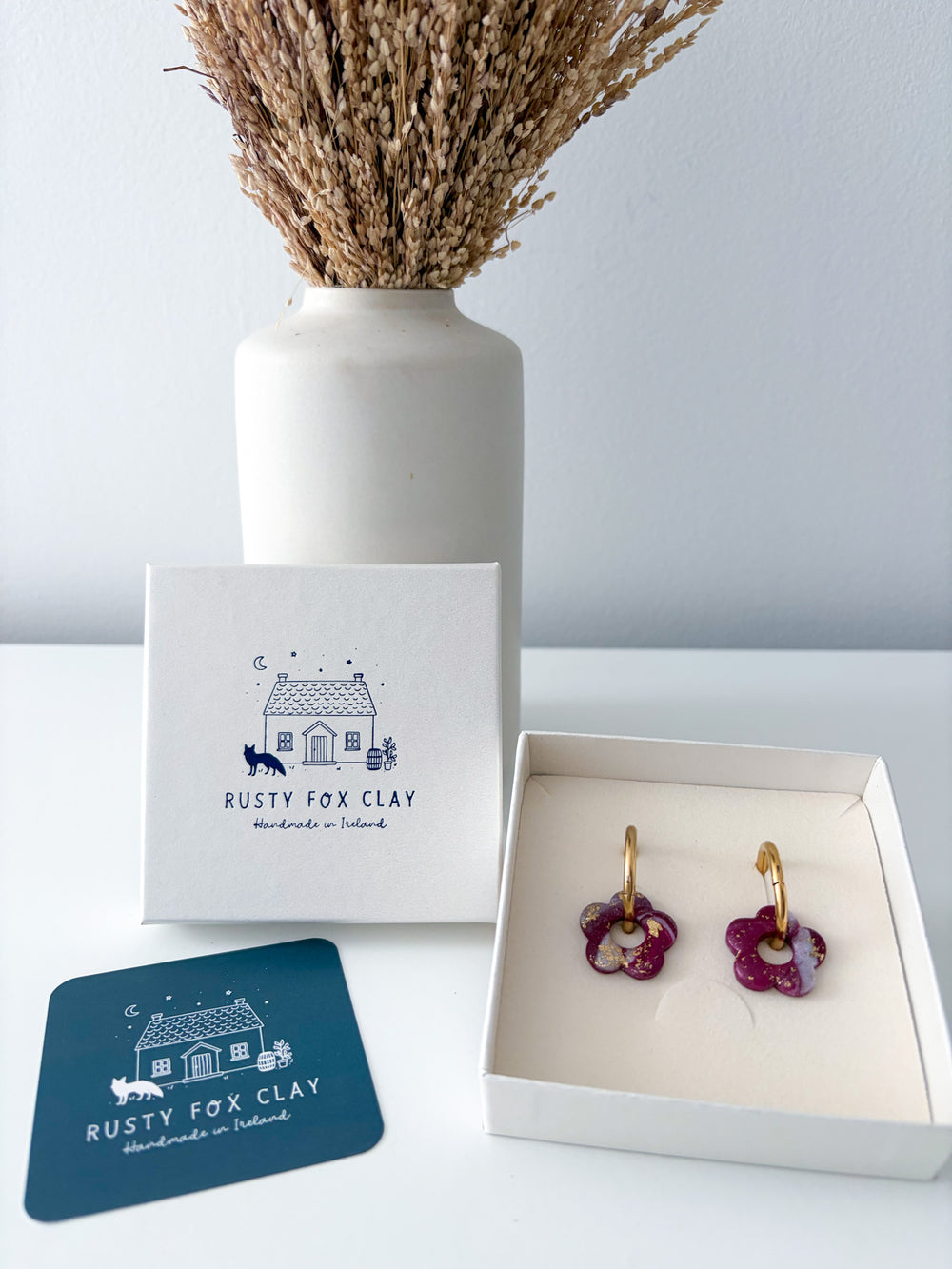 Rusty Fox Clay handmade earrings in a floral design, featuring deep magenta petals and gold-tone hooks, displayed in an open white box next to its branded packaging and a blue coaster.