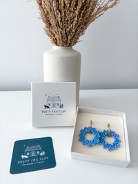 Blue sun-shaped hoop earrings with gold hooks displayed in an open white gift box next to a branded Rusty Fox Clay card and box, with a white vase of dried grass in the background.