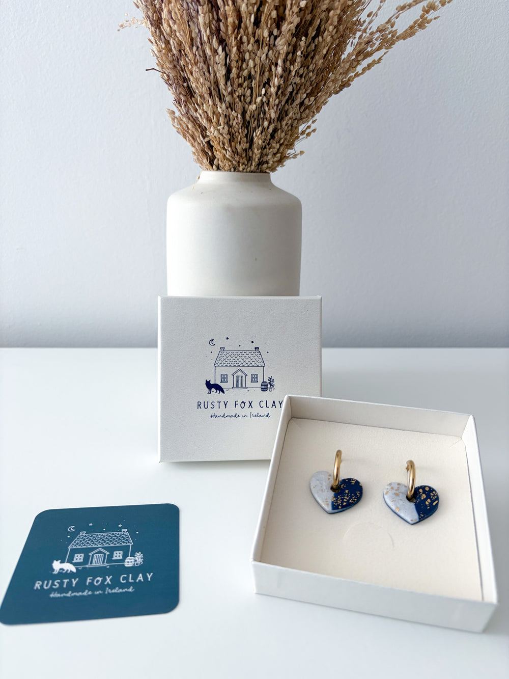 Heart-shaped earrings with blue and white enamel and gold-tone hooks, displayed in an open white gift box next to a branded card and a white vase with dried grass on a light gray background.
