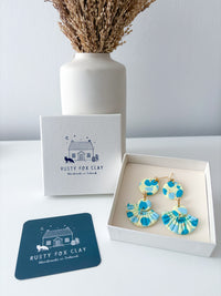 Blue and yellow patterned dangle earrings displayed in an open white gift box next to a branded Rusty Fox Clay box and a dark teal branded card, with a white vase of dried grass in the background.
