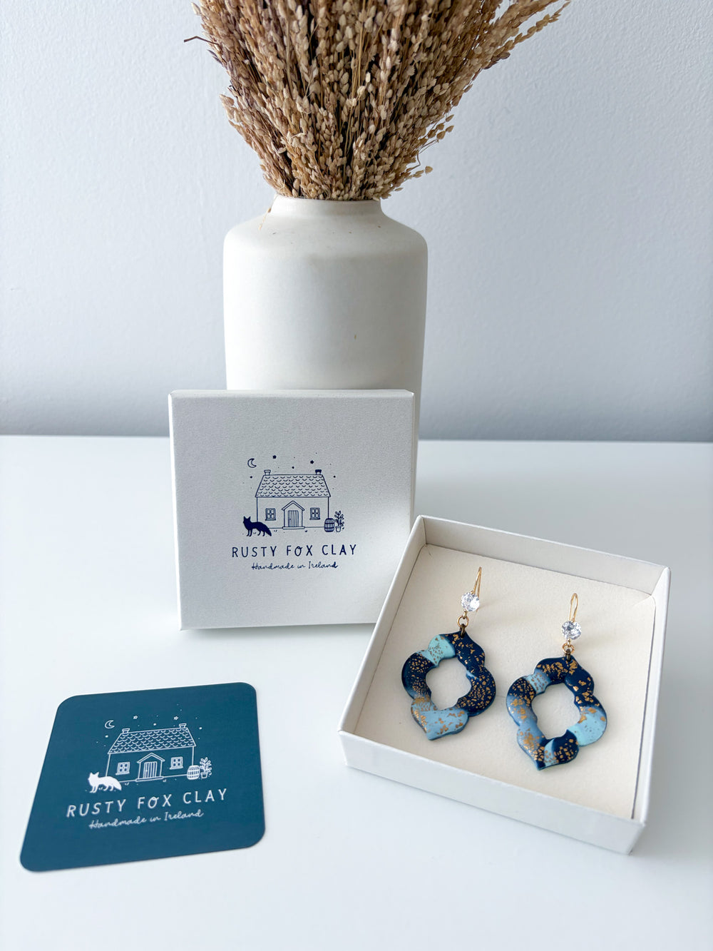 Rusty Fox Clay handmade earrings displayed in an open white gift box next to the brand's white box and a teal brand card, with a white vase of dried grass in the background.
